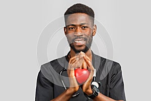 Young professional african doctor cardiologist with stethoscope holding red heart in hand, standing against gray background.