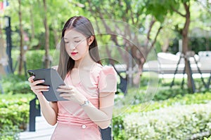 Young professional Asian business woman with long hair is smiling in the garden while looking at the tablet in her hand, work from