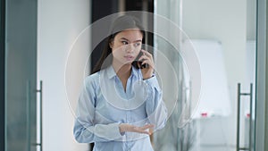 Young professional asian woman manager talking to client or partner via cellphone, standing at modern office corridor