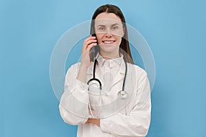 Young professional doctor female in white medical uniform and stethoscope talking on mobile phone, posing isolated over blue