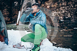 Young professional male tourist is drinking in the mountains near the river in winter.