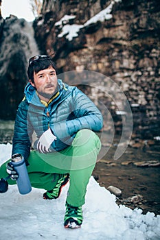 Young professional male tourist is drinking in the mountains near the river in winter.