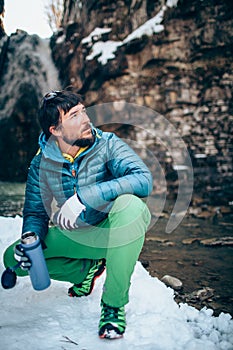 Young professional male tourist is drinking in the mountains near the river in winter.