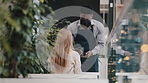 Young professional male waiter in apron restaurant worker in medical mask serves client in cafe brings coffee in paper