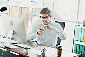 Frustrated businessman angrily yelling at smartphone while working on computer in stylish modern office setting.