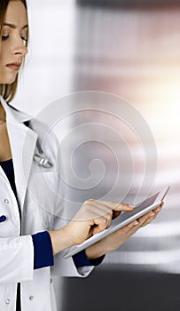 Young professional woman-doctor is using a tablet computer, while standing in a clinic. Portrait of beautiful female