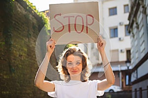 Young protesting woman in white shirt and jeans holds protest sign broadsheet placard with slogan `Stop` for public demonstratio