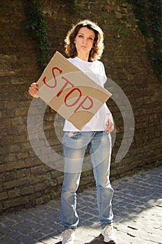 Young protesting woman in white shirt and jeans holds protest sign broadsheet placard with slogan `Stop` for public demonstratio