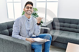 Young psychologist man at consultation office pointing to the back behind with hand and thumbs up, smiling confident