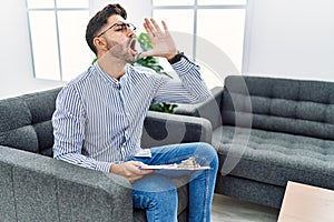 Young psychologist man at consultation office shouting and screaming loud to side with hand on mouth