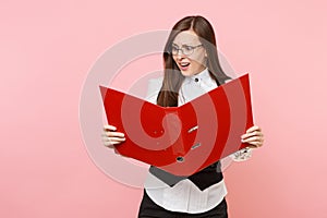 Young puzzled business woman in suit, glasses looking on red folder for papers document on pastel pink