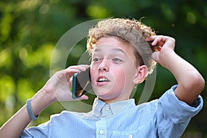 Young puzzled child boy talking on cellphone outdoors in summer park