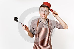 Young puzzled fun man chef or waiter in striped brown apron, shirt holding red empty stewpan black ladle isolated on