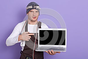 Young radioman wears white casual shirt, cap and apron, poins at blank screen of laptop over lilac background, radiotrician has
