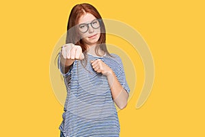 Young read head woman wearing casual clothes and glasses punching fist to fight, aggressive and angry attack, threat and violence