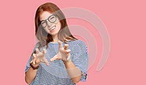 Young read head woman wearing casual clothes and glasses smiling funny doing claw gesture as cat, aggressive and sexy expression