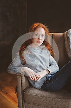 Young readhead woman relaxing at home in cozy chair, dressed in casual sweater and jeans