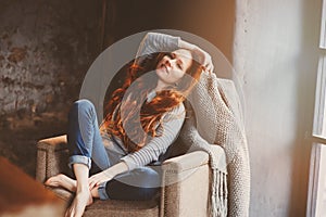 Young readhead woman relaxing at home in cozy chair, dressed in casual sweater and jeans