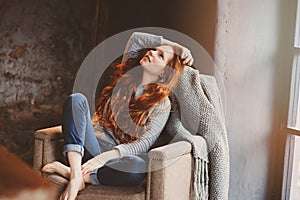 Young readhead woman relaxing at home in cozy chair, dressed in casual sweater and jeans