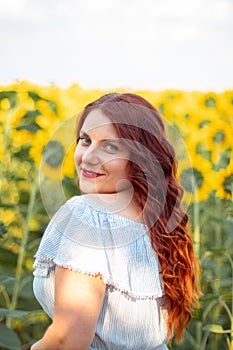 Young red hair Caucasian girl in summer cotton dress in a field of yellow sunflowers on a sunny day