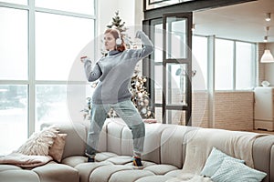 Young red-haired beaming girl in a grey sweater dancing on a sofa