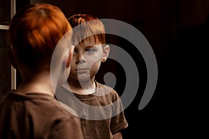 A young red-haired boy looks thoughtfully at himself in the mirror on a dark background. Childhood crisis, depression concept.