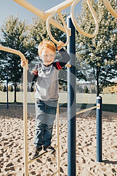 Young red-haired Caucasian boy hanging on monkey bars in park on playground. Summer outdoors activity for kids. Active preschool