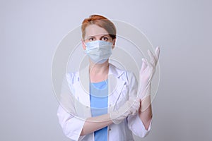 Young red-haired female medic in white coat and protective medical mask pulls a latex rubber glove over her hand