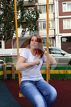 A young red-haired girl with freckles on her face in a white t-shirt and blue jeans rides on a swing.