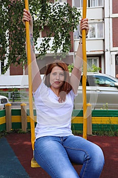 A young red-haired girl with freckles on her face in a white t-shirt and blue jeans rides on a swing.