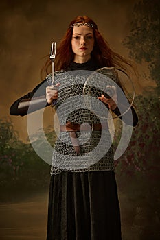 Young red haired girl-knight posing holding serving fork and lid like sword and shield against vintage studio background