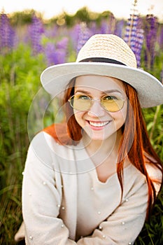 Young red haired girl with long hair in a wicker hat with a ribbon. Woman sitting with on a field of lupins at sunset and smiling