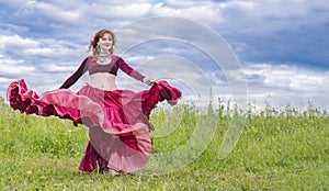 Young red-haired girl in red dance dress dances on green meadow against blue summer sky.