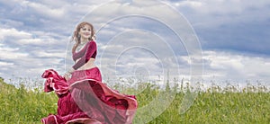 Young red-haired girl in red dance dress dances on green meadow against blue summer sky.