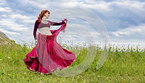 Young red-haired girl in red dance dress dances on green meadow against blue summer sky.