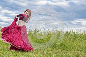 Young red-haired girl in red dance dress dances on green meadow against blue summer sky.