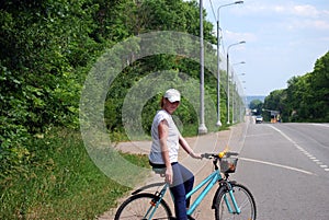 The young red-haired girl in a white cap and a white t-shirt stands on a roadside with bicycle, in the summer sunny day.