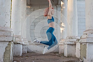 Young red-haired sportswoman jumping in the air. In a turquoise tracksuit, leggings, against the background of a concrete white