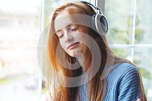 Young red haired woman with headphones listening to music at home, relaxing, enjoying songs, closed eyes, soul music