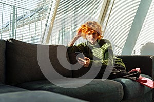 young red-haired woman in living room on sofa using phone
