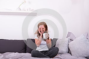 Young, red-haired woman sitting on the bed and enjoying her coffee