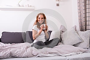 Young, red-haired woman sitting on the bed and enjoying her coffee