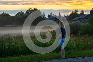 A young red-haired woman on a summer evening looks at the rural landscape. Fog over a field in the village. Shallow depth of field