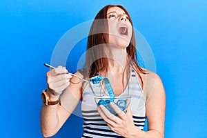 Young red head girl eating measure meter doing diet for weight loss angry and mad screaming frustrated and furious, shouting with