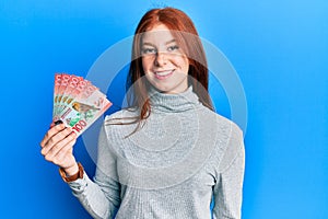 Young red head girl holding 100 new zealand dollars banknote looking positive and happy standing and smiling with a confident