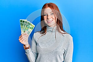 Young red head girl holding 50 israel shekels banknotes looking positive and happy standing and smiling with a confident smile