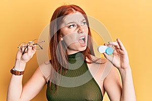 Young red head girl holding glasses and contact lenses angry and mad screaming frustrated and furious, shouting with anger