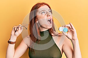 Young red head girl holding glasses and contact lenses angry and mad screaming frustrated and furious, shouting with anger looking