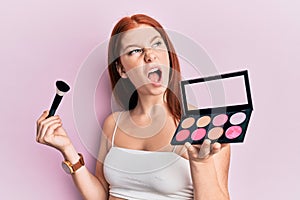Young red head girl holding makeup brush and blush palette angry and mad screaming frustrated and furious, shouting with anger