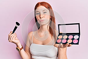 Young red head girl holding makeup brush and blush palette looking at the camera blowing a kiss being lovely and sexy